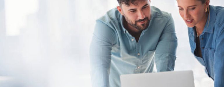 Businessman and businesswoman working on a laptop computer. They are wearing matching casual clothes that could be a uniform. They are standing in a board room with a window behind them