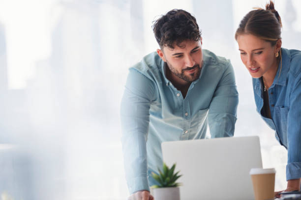 Businessman and businesswoman working on a laptop computer. They are wearing matching casual clothes that could be a uniform. They are standing in a board room with a window behind them